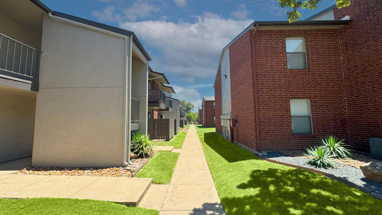 Apartment building with balconies, fenced patios, and a stairwell, surrounded by green grass and a clear blue sky.