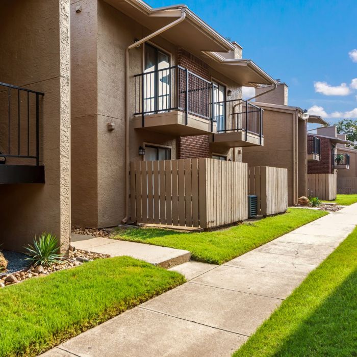 Apartment building with balconies, fenced patios, and a stairwell, surrounded by green grass and a clear blue sky.