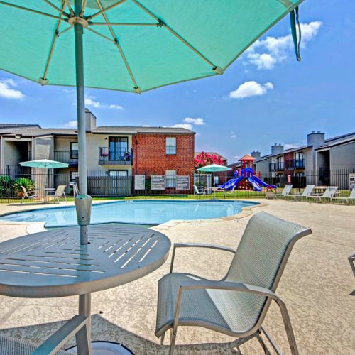 Poolside area with umbrella table and chairs near apartment buildings, playground visible in the background.