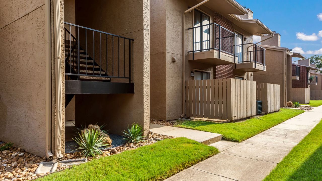 Apartment building with balconies, fenced patios, and a stairwell, surrounded by green grass and a clear blue sky.