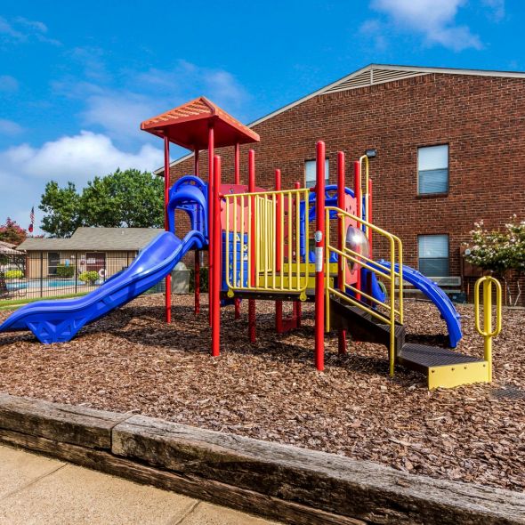 Playground with colorful slides and climbing structures on wood chips, next to a brick building under a clear sky.