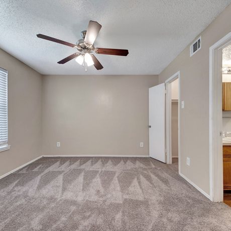 Empty room with beige walls, carpeted floor, ceiling fan, window with blinds, and an open door to a bathroom.
