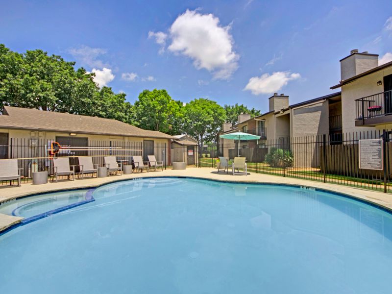 Outdoor pool area with lounge chairs and umbrella, surrounded by buildings and trees under a partly cloudy sky.