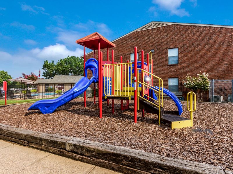 Playground with colorful slides and climbing structures on wood chips, next to a brick building under a clear sky.